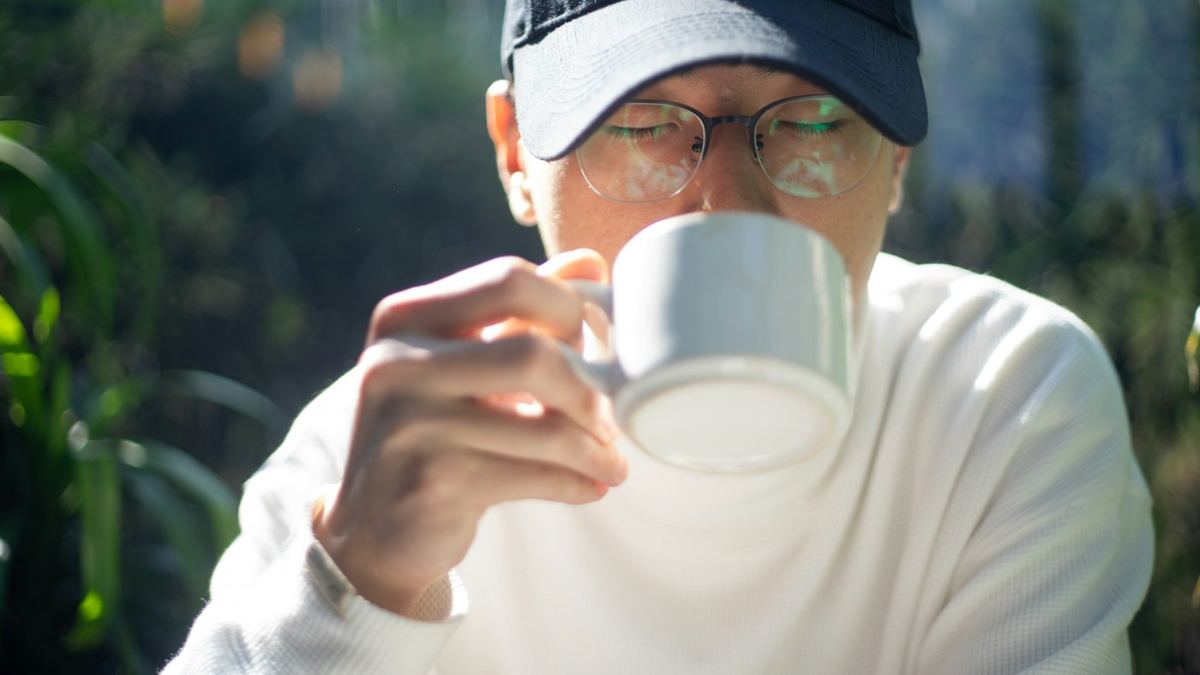 Man med keps och glasögon som dricker ur en vit kopp utomhus