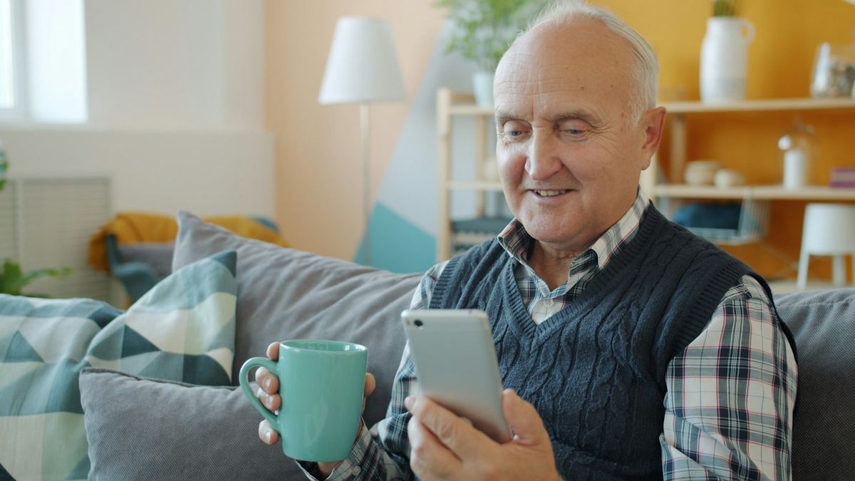 Leende äldre man i soffan med en kopp te i ena handen och mobilen i den andra